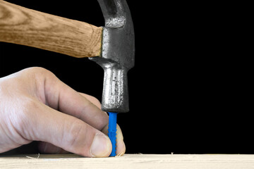 A worker's hand with an old rusted hammer hammers a plastic dowel into a hole into a light wood surface isolated on a black background with space for text.