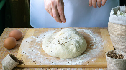 The hands of an elderly woman knead the dough for bread on a wooden board on the table, on the table there are 2 eggs, there are linen bags with spices cumin and coriander and flour. World Bread Day