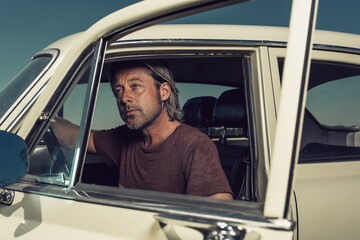 Blond man in t-shirt sitting in classic car with open door.