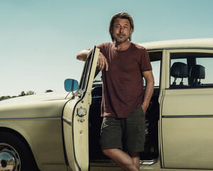 Blond man in brown t-shirt and shorts standing by open door of a classic car on a sunny day in the...