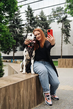 Young reddish woman at the street with her dalmatian dog taking a picture with mobile phone. Lifestyle outdoors with pets. Love, care and friendship