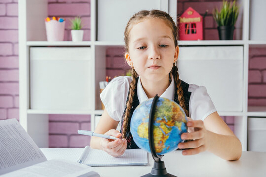 Little schoolgirl girl sits at the desk, looks at the globe and does her geography homework