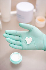 Heart shaped cream on the palm of a gloved hand and a jar with cream on the background of skin care products.