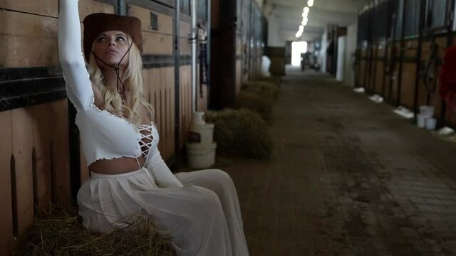sexy blonde woman with traditional cowboy hat is sitting alone in barn