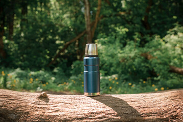 Traveling blue thermos on a log in the forest. Outdoor camping reusable flask. Concept of hiking, camping in nature - obrazy, fototapety, plakaty