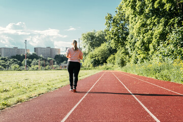 Training at the stadium, outdoors. Young plus size woman walking outside on sunny summer day. Overweight woman doing sports, back view. Active lifestyle, weight loss