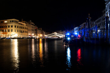 Night view of the main bridge of Venice ("Ponte di Rialto"), with a boat in the frontground and the city lights reflected on the water.