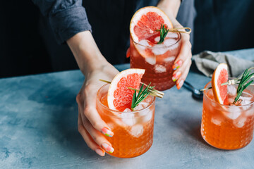 Female bartender hand holding grapefruit cocktail lemonade glasses  with ice and rosemary. - obrazy, fototapety, plakaty