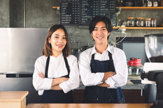 Couple asian barista owner coffee shop standing at counter welcome the customer. Cheerful two young barista man and woman in apron serving cup of hot coffee in cafe.