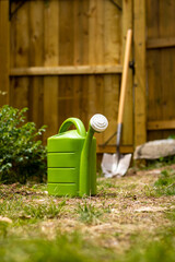 A simple close-up shot of a green watering can with a large shovel and wooden fence in the background.