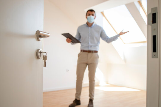 Portrait Of Male Real Estate Agent With Face Mask Standing In New House During Corona Virus Pandemic.