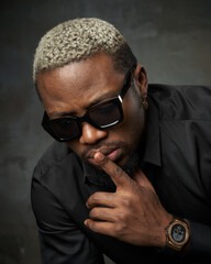 Close-up face studio portrait of young stylish glamor handsome african man with short white hair in black silk shirt and sunglasses posing against grey concrete wall