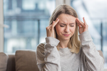 A woman holding her head anxious about an unbearable headache, sitting at home on the couch
