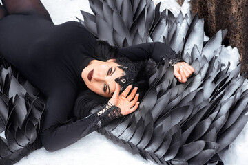 portrait of a beautiful young brunette woman dressed as a black angel, who lies on the snow and looks at the camera