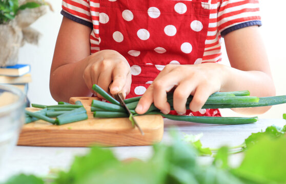 The concept of proper nutrition from an early age. Children's hands are slicing green onions on a cutting board against a background of various vegetables, side view