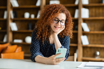 Smiling cheerful beautiful caucasian woman in eyeglasses with curly hairstyle sitting at desk and using smartphone, charming female office employee chatting on the phone online, messaging