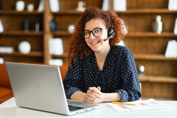 Young curly woman wearing wireless headset using a laptop computer for communication with customers...