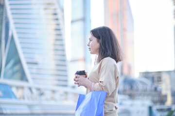 beautiful young woman enjoying a cup of coffee, against the backdrop of skyscrapers Smiling hipster girl with cup of hot beverage on city street.