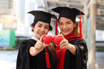 Two happy smiling graduated students holding small red heart gift and giving to camera, young beautiful Asian women on their commencement day, people celebrating successful education on graduation day