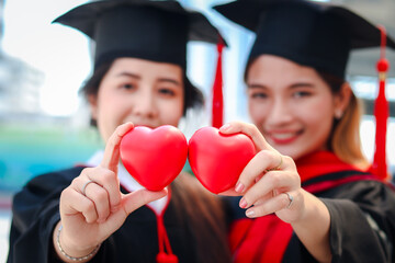 Two happy smiling graduated students holding small red heart gift and giving to camera, young...