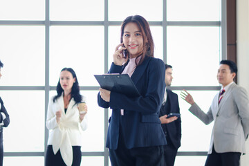 Smart and beautiful young businesswoman with black long hair in suit talking on mobile phone at the window office workplace with businesspeople colleague as blurred background.