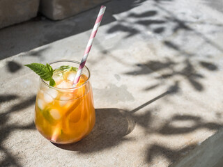 Cold orange cocktail with raspberry leaves, ice. Cocktail in glass with cocktail straw.  The background is a natural concrete block with cement. Sunshine, shadow from tree foliage