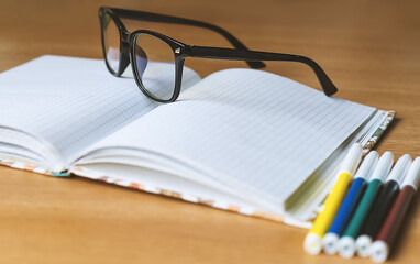 On the table is an office notebook, glasses, markers. Office supplies. Black-rimmed glasses, colored markers, and an open notebook lie on the table in close-up. Selective Focus.