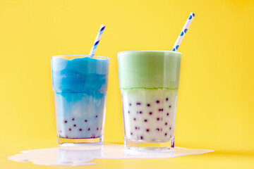 a glass of green matcha latte and blue matcha latte with tapioca balls, spilled milk on a yellow background