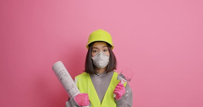Reapair building and decorating concept. Pensive Asian woman builder concentrated above considers future architectural plan holds blueprint and brush wears protective helmet respirator uniform
