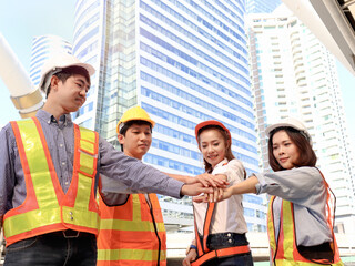 Group of four young Asian male and female engineers wearing safety vest and helmet, putting hands join together at outside office in downtown city, partnership colleagues holding hands as commitment o