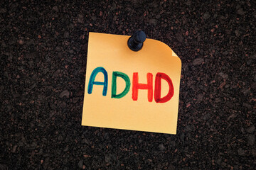 The abbreviation ADHD written on a yellow paper note pinned to a cork board
