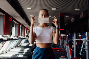 Fototapeta na wymiar A fit woman in a face mask to avoid the spread of coronavirus is doing bicep curls with dumbbells. A sporty girl in a surgical mask is posing during the arms workout in the gym.