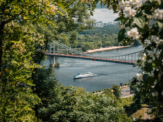 The boat floats on the river under the pedestrian bridge in Kiev. The Dnieper River and the green Trukhaniv island
