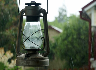 An old kerosene / oil lamp hangs in the rain amid green plants and village houses.