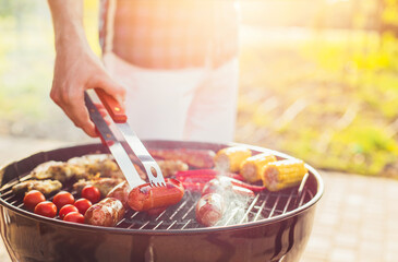 Man fries meat and vegetables on the grill, flips sausages with tongs, corn cobs, chili peppers and chicken wings outdoors