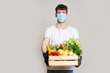 Food delivery concept during quarantine. A man in a mask, gloves and a white T-shirt holds a box of fresh food in his hands. Food delivery from the supermarket, from farmers home.