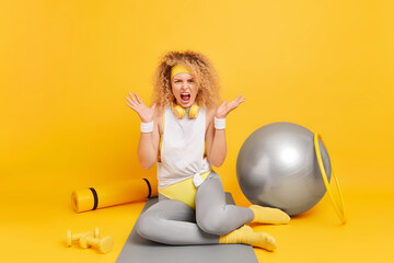 Indoor shot of angry curly haired European woman screams loudly spreads palms expresses negative emotions feels irritated after hard fitness training with dumbbells hula hoop and swiss ball.