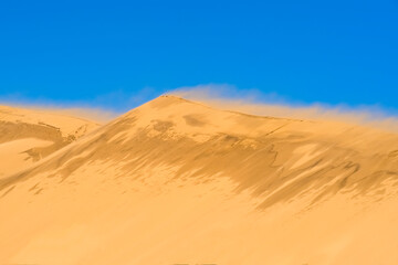 Fototapeta na wymiar Dune against the background of a bright blue sky. The wind blows the sand off the ridge of the dune. Wild nature landscape. Desktop wallpaper