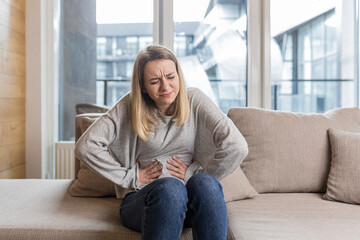Young woman sitting on sofa holding hands on stomach twisted from pain, poisoned or menstrual pain, woman at home