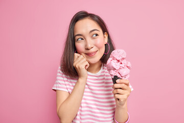 Photo of pretty Asian teenage girl looks dreamily aside recalls pleasant memories eats tasty summer dessert holds big cone ice cream dressed in casual t shirt models against pink background. - obrazy, fototapety, plakaty