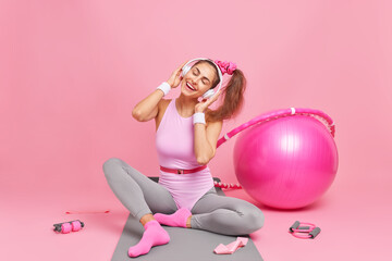 Obraz na płótnie Canvas Attractive cheerful sportswoman tilts head enjoys favorite melody dressed in active wear leads healthy active lifestyle does gymnastics or aerobics isolated over pink background. Home workout