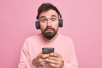 Indoor shot of shocked bearded man stares bugged eyes at camera uses mobile phone listens audio track via wireless headphones dressed in casual clothing isolated over pink background. Perfect sound