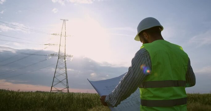 A power engineer stands in front of a high-voltage power line, holds paper drawings in his hands and looks at them as the sun goes down.