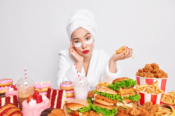 Serious young Asian woman looks directly at camera holds french fries poses near table full of harmful food applies patches under eyes wears bathrobe soft towel poses against white background