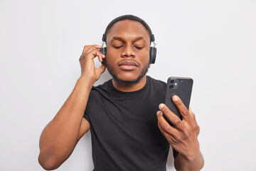 Waist up shot dark skinned man with beard looks attentively at smartphone device chooses song from playlist uses stereo headphones wears black t shirt isolated over white background. Listening music