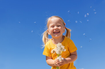 A little girl with a bunch of white dandelions against blue sky. Smiling child in a yellow t-shirt,...