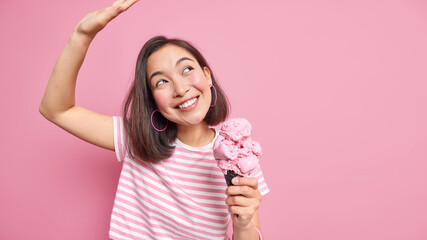 Optimistic Asian woman dances carefree keeps arm raised up holds appetizing ice cream eats high calorie snack dressed in striped t shirt isolated over pink background copy space for your promo