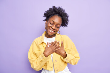 Thank you from bottom of heart. Pleased black woman makes thankful gesture apprectiates something tilts head smiles gently keeps eyes closed wears yellow jacket isolated over purple background