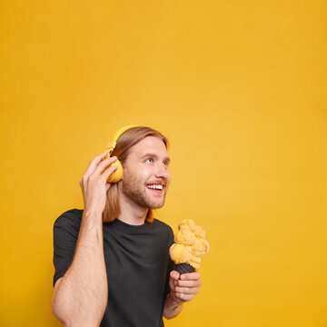 Vertical image of cheerful guy focused above with glad expression eats delicious ice cream during summer time listens music via wireless headphones stands against yellow background copy space