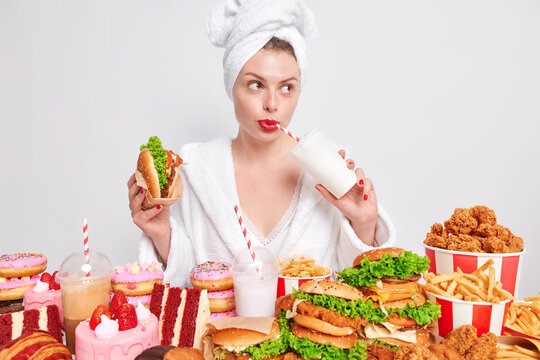Pensive European lady with red painted lips drinks soda eats burger looks away addicted to fast food isolated over white background thinks about something. Cheat meal unhealthy nutrition concept